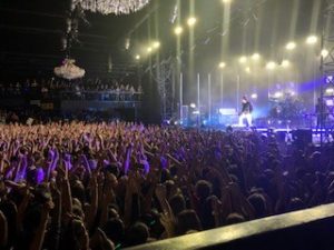 Band 5 Seconds of Summer on stage, the audience takes up most of the picture, the band is lit by white and blue lights