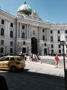 This photo was taken in Vienna and shows a beautiful white building. It has people in the photo as well as a taxi car. 