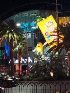 A statue of the yellow m&m holding up a big box of m&ms at night.